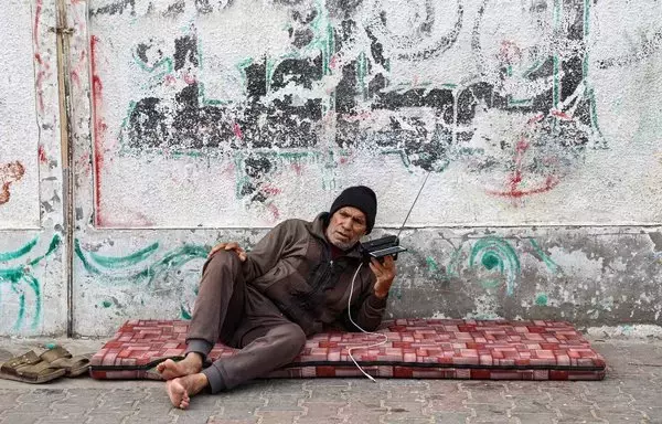 رجل فلسطيني يستمع إلى جهاز الراديو الخاص به في مخيم للاجئين في رفح بجنوب قطاع غزة في 17 كانون الأول/ديسمبر، وسط استمرار المعارك بين إسرائيل وحماس. وفي قطاع غزة أصبح من المستحيل الآن توصيل أجهزة الكمبيوتر أو أجهزة التلفزيون أو إعادة شحن الهواتف أو الوصول إلى الإنترنت من دون مولدات أو ألواح شمسية، وهي رفاهية لا يستطيع سوى عدد قليل من سكان غزة تحملها. [محمد عابد/وكالة الصحافة الفرنسية]